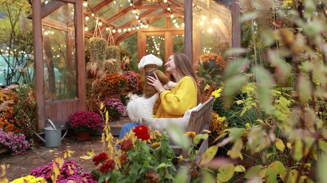 Happy girl pet owner holding a small white dog in a cozy fall garden filled with potted flowers. Warm autumn evening landscape with glowing greenhouse lights, backyard, pet friendship concept.