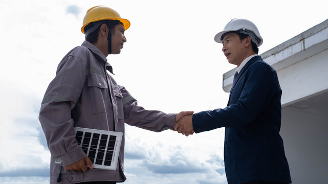 Engineer and construction worker shaking hands at a building site while holding a solar panel, representing renewable energy, project partnership, sustainable construction and clean energy solutions.