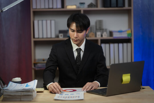 Asian Businessman Working Late Night in Office, Serious Executive Reading Financial Paperwork and Analyzing Reports at Desk