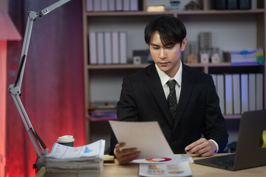 Asian Businessman Working Late Night in Office, Serious Executive Reading Financial Paperwork and Analyzing Reports at Desk