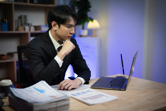 Asian Businessman Working Late Night in Office, Serious Executive Reading Financial Paperwork and Analyzing Reports at Desk