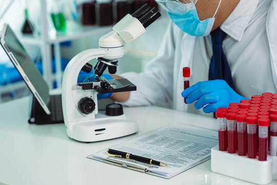 Scientist Holding Blood Sample Test Tube with Microscope and Rack of Blood Vials in Medical Laboratory