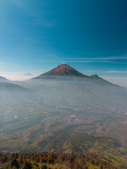Fototapeta premium A stunning aerial view of Mount Sindoro with the Seroja hills in the foreground, located in Indonesia. An aerial photo of the mountain taken in the late morning against a blue sky.