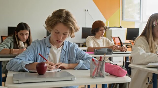 Caucasian teenage boy sitting at desk in classroom, pushing hair back, and writing in notebook. Clip is ideal for school marketing or educational software promos