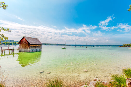 sunny day at Lake Ammersee in Bavaria, Germany, Europe. rustic wooden boathouse on stilts over calm, clear turquoise water. sailboats and yachts are anchored across the calm lake under bright sky