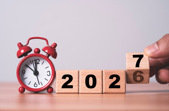 Hand turns wooden blocks from 2026 to 2027 beside an alarm clock, representing New Year countdown, time transition, deadline, planning, fresh start and preparation for the year ahead.