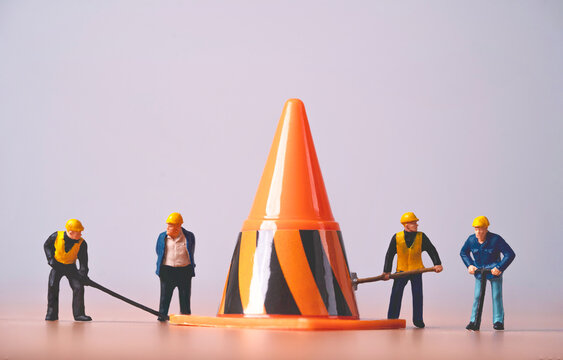 Miniature construction workers surround a large traffic cone, symbolizing roadwork, workplace safety, maintenance, teamwork, hazard control and infrastructure repair on a work site.