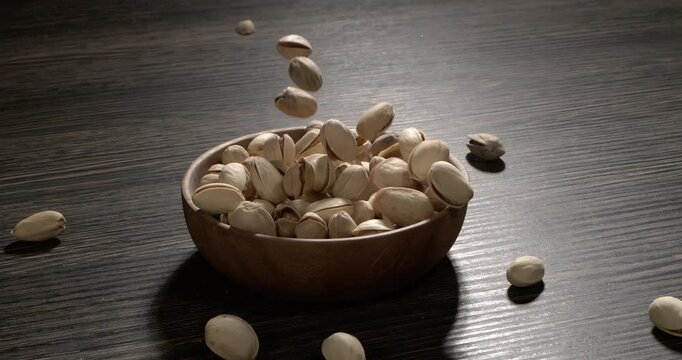 Wooden bowl filled with pistachios on dark wooden table, tasty salted nuts scattered around, healthy snack and natural food concept. Shot on super slow motion camera 1600 fps.