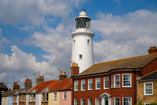 Southwold Lighthouse rises above rows of pastel and brick terraced houses in Suffolk, its white tower and lantern room set against a bright blue sky with billowing clouds.