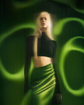 Blonde woman in black crop top and shiny green skirt behind vertical distorted glass bars on abstract green and black background