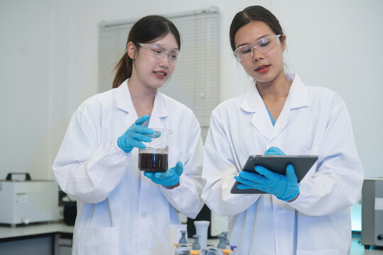 Two Asian Female Scientists Analyzing Dark Liquid Sample with Tablet in Modern Laboratory