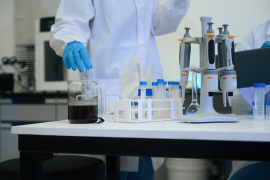 Close up of Scientist in White Lab Coat and Blue Gloves Stirring Dark Liquid Sample in Beaker with Laboratory Equipment