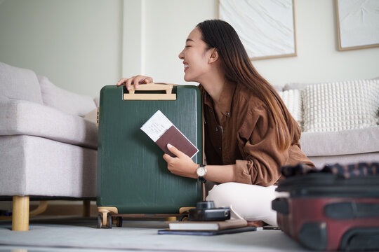 Asian traveler holding a passport and ticket beside her packed suitcase.
