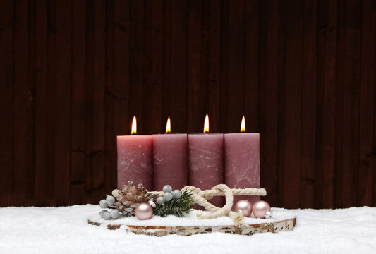 Fourth Sunday of Advent: A rustic Advent wreath in the snow with four pink-flame pillar candles on a tree stump. In the background, a wooden wall with a copy room.
