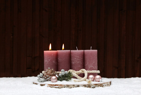 Second Sunday of Advent: A rustic Advent wreath in the snow with two pink-flame pillar candles on a tree stump. In the background, a wooden wall with a copy room.
