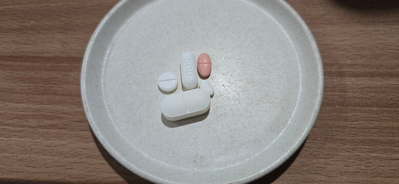 Various types of pills are placed on a white plate, including large white oval, white oval printed with the number 500, white round pills, pink capsule-shaped pills. Medication to be taken after meals