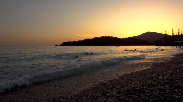 The setting sun casts golden light on the seashore of a tropical resort. Silhouettes of people swimming are visible in the ocean waves. 4k video.