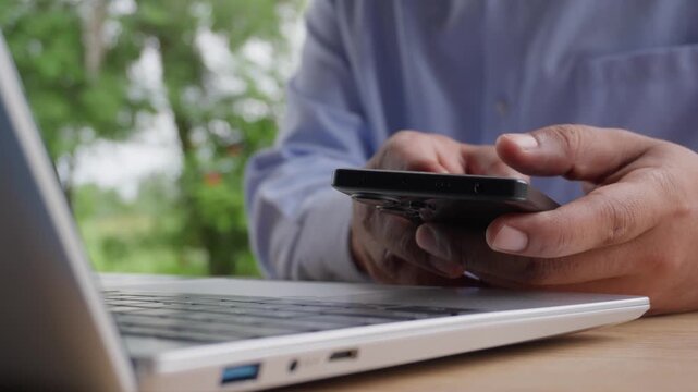 Person using mobile smartphone on office wooden desk with laptop computer in foreground and scenic green nature trees background, concept of remote work lifestyle and daily mobile technology use.