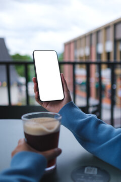 Vertical mockup image of a woman holding and using mobile phone with blank desktop screen while drinking coffee in cafe