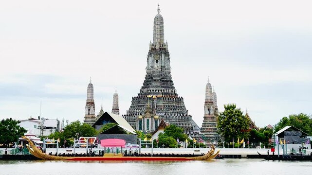 A golden Thai royal barge processional rowing past the iconic Wat Arun temple on the Chao Phraya River in Bangkok, Thailand during a traditional cultural ceremony under a bright daylight sky.