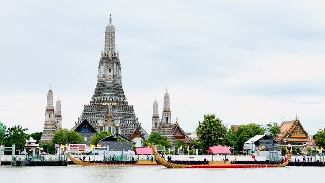 A golden Thai royal barge processional rowing past the iconic Wat Arun temple on the Chao Phraya River in Bangkok, Thailand during a traditional cultural ceremony under a bright daylight sky.