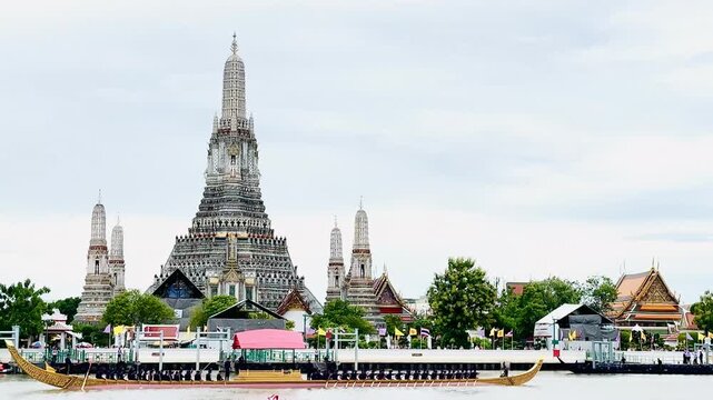A golden Thai royal barge processional rowing past the iconic Wat Arun temple on the Chao Phraya River in Bangkok, Thailand during a traditional cultural ceremony under a bright daylight sky.
