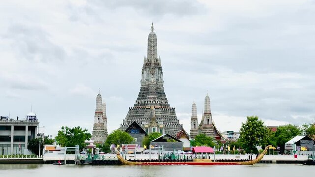 A golden Thai royal barge processional rowing past the iconic Wat Arun temple on the Chao Phraya River in Bangkok, Thailand during a traditional cultural ceremony under a bright daylight sky.