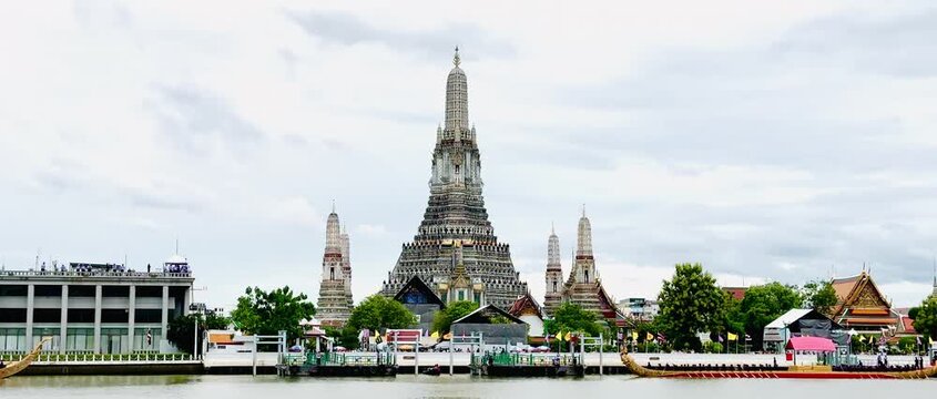A golden Thai royal barge processional rowing past the iconic Wat Arun temple on the Chao Phraya River in Bangkok, Thailand during a traditional cultural ceremony under a bright daylight sky.