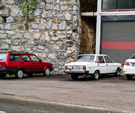 A red Tofas Kartal station wagon and a white Tofaş Murat 131 classic car are parked in front of a natural stone wall along a road in Anatolia; these are classic Turkish cult cars from the 1980s 5.4.25