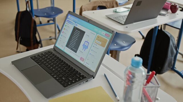 Laptop displaying educational coding software sitting on school desk in empty classroom, high angle shot. Clip is ideal for promoting STEM education, e-learning platforms, and school technology