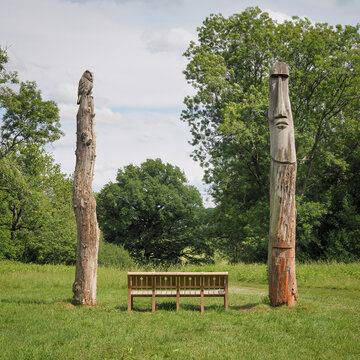 Two tall carved wooden totem sculptures depicting an owl and a face frame a park bench along the woodland trail in Bedwyn Brail, Wiltshire, UK