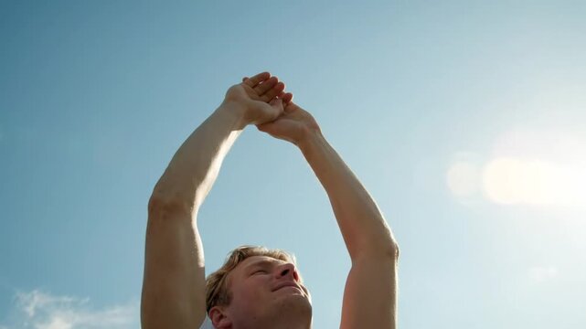 A young man stands under a bright sun with eyes closed and arms raised, finding peace and tranquility. He clasps his hands above his head in a meditative gesture against a clear blue sky.