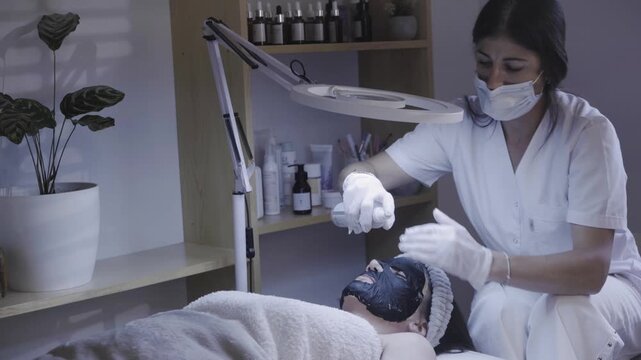 Gloved esthetician massaging dark clay mask onto client face