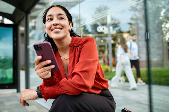 Smiling young woman using smartphone at city bus stop