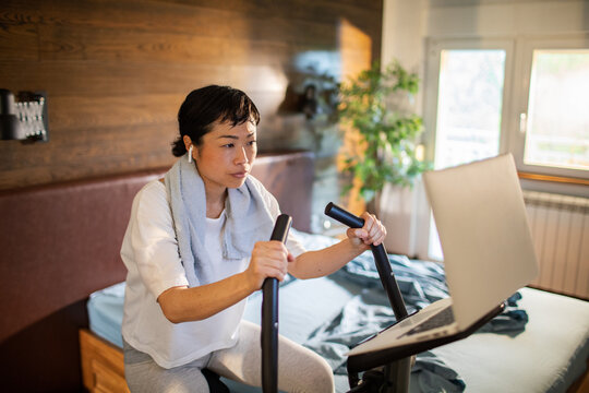 Woman on exercise bike following workout on laptop in home bedroom