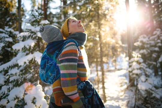 Mother carrying toddler son on back while enjoying snow in winter forest