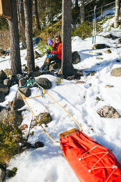 Red pulk sled on snowy landscape near rocks and woman sitting with child near tree