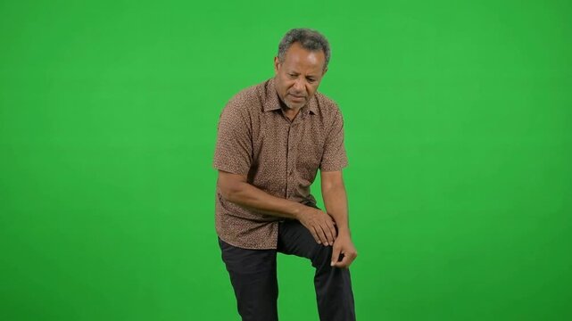 Elderly man wearing a patterned shirt kneels on a bright green background, expressing concern for his knee