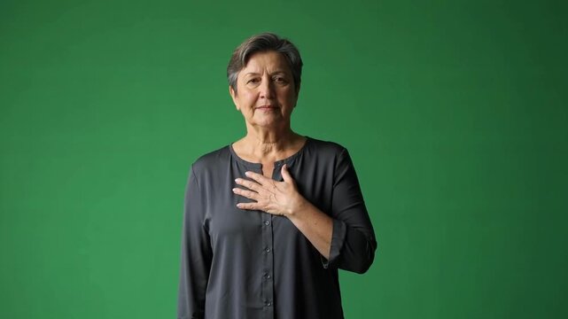 Portrait of a mature woman with short hair and a gray blouse standing against a green background, hand on chest expressing sincerity