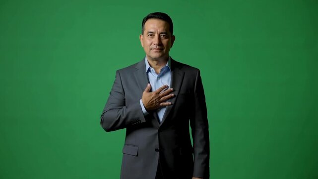 A middle-aged man in a formal suit with a green background, expressing sincerity and confidence by placing his hand on his chest