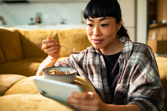 Woman eating oatmeal while watching tablet on sofa at home