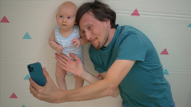 Happy father showing baby on phone beside soft mat.