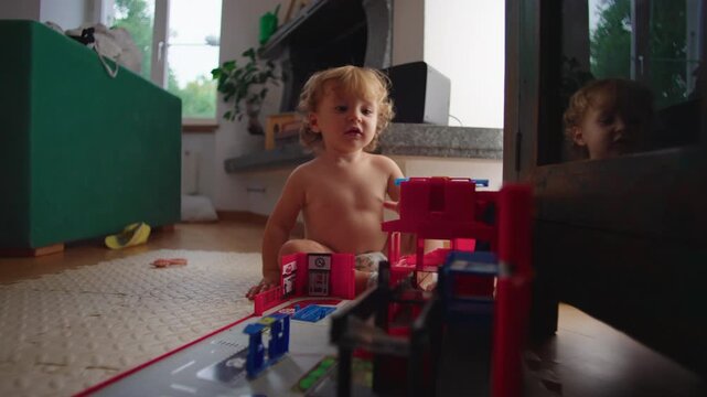 Toddler sitting on floor while opening toy fire station playset indoors capturing curiosity, hands on learning and authentic everyday childhood imagination at home