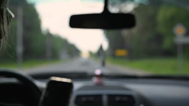 Blurred view car interior with the driver hands on the steering wheel and trees lining the road ahead. Other cars are visible in the distance as the vehicle moves along a clear street