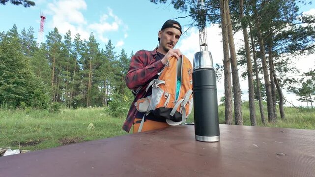 Seated man wearing backward cap and red plaid shirt pours beverage from tall black metal thermos into small cup on wooden picnic table while orange backpack rests beside him, hands steady thermos