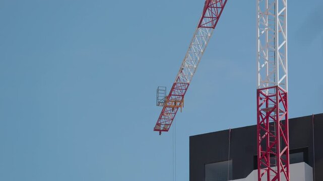 Red and white tower crane jib slowly lowering next to a modern building on a clear day
