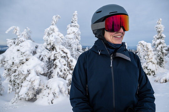 Smiling mature woman wearing ski goggles and helmet standing in front of snow covered trees in winter