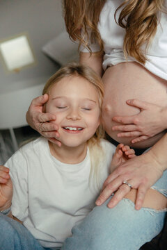 Smiling girl resting her head on pregnant mother's belly for listening to heartbeat at home