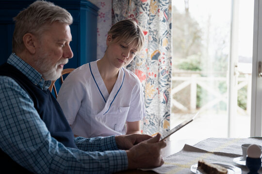 Female healthcare worker sitting with senior man reading news on tablet PC while sitting at table in home