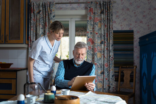 Female caregiver standing beside elderly man using digital tablet while sitting for breakfast at home